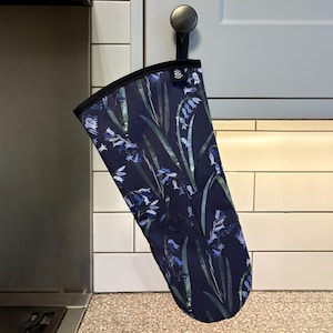 May include: A blue oven glove with a floral pattern of bluebells and green leaves. The glove is hanging from a hook on a cabinet.