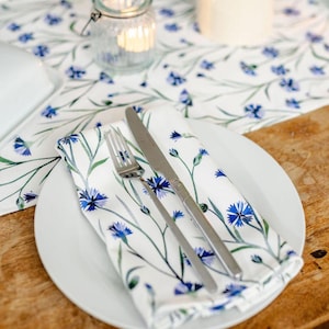 Pode incluir: Um prato branco com uma toalha de mesa dobrada, garfo e faca. A toalha de mesa e o caminho de mesa apresentam um padrão floral azul. Uma vela acesa e um frasco de vidro com uma vela estão no fundo. A mesa é de madeira.