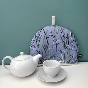 May include: A white ceramic teapot, teacup, and saucer set next to a blue floral tea cozy. The tea cozy features a bluebell pattern on a light blue background. The teapot has a rounded shape with a handle and spout.