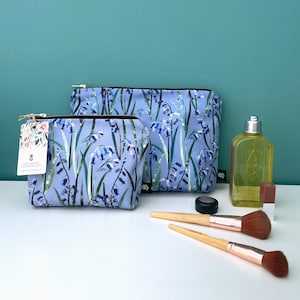 Puede incluir: Dos bolsas de maquillaje con cremallera de estampado floral azul. La bolsa más grande está en el fondo y la más pequeña en primer plano. Las bolsas están sobre una superficie blanca con un compacto marrón, dos pinceles de maquillaje y una botella transparente de líquido.