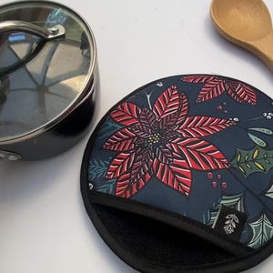Könnte beinhalten: Ein runder, blauer Topflappen mit einem roten und grünen Weihnachtsstern-Design. Der Topflappen hat einen schwarzen Rand und eine schwarze Stoffschlaufe auf der Rückseite.