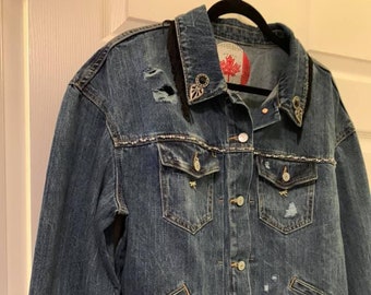 Peut inclure: Veste en jean délavé avec des détails déchirés, une bordure en franges noires et des ornements décoratifs. La veste a une fermeture boutonnée, des poches poitrine et un écusson du drapeau canadien au dos.