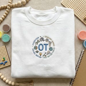 Könnte beinhalten: Weißes Sweatshirt mit Rundhalsausschnitt und gesticktem Design. Das Design zeigt die Buchstaben "OT" in Blau, umgeben von Illustrationen und Texten, die sich auf Ergotherapie beziehen, darunter "Cognition" und "Sensory Rings".