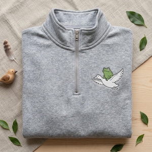 May include: A grey quarter-zip sweatshirt featuring an embroidered design of a green frog riding a white bird. The sweatshirt has a silver zip and a folded collar. The design is on the left chest.