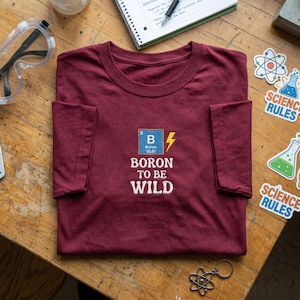 May include: A maroon short-sleeve t-shirt featuring an embroidered blue square with the chemical symbol B for Boron, the number 5, and the atomic weight 10.81, next to a yellow lightning bolt, with the white embroidered text Boron To Be Wild.
