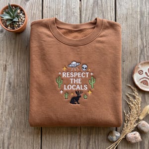 May include: A brown crewneck sweatshirt featuring an embroidered design with the text "RESPECT THE LOCALS" in white. The graphic includes a grey UFO, an alien head, a black jackalope, green cacti, and small yellow stars.