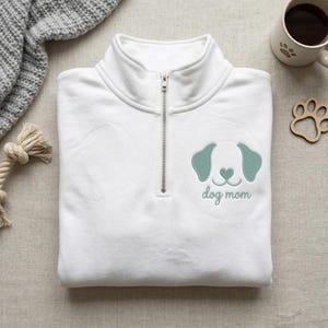 May include: White quarter-zip sweatshirt with a teal dog face design and the words "dog mom" embroidered below. The sweatshirt has a silver zipper and is folded neatly. A mug and dog paw-shaped coaster are in the background.