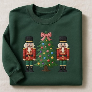 May include: A dark green sweatshirt with an embroidered design of two nutcrackers and a Christmas tree. The nutcrackers wear red and green uniforms with black hats. The tree is decorated with colourful baubles and a pink bow.