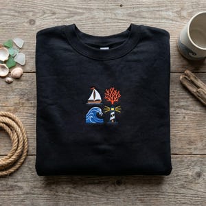 Könnte beinhalten: Schwarzes Sweatshirt mit Rundhalsausschnitt und maritimen Stickereien. Das Design umfasst ein Segelboot, Korallen, eine Welle und einen Leuchtturm. Das Sweatshirt ist aus weichem Material und ideal für Meeresliebhaber.