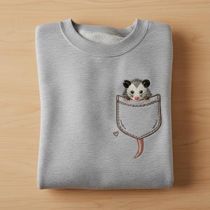 May include: A pale grey sweatshirt with an embroidered pocket design. The pocket features a cute opossum peeking out, with a small heart detail. The opossum has black ears, a grey body, and a pink nose and tail.