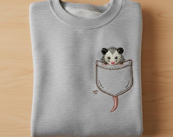 May include: A light gray sweatshirt with a detailed embroidered pocket design. The pocket features a cute opossum peeking out, with a small heart detail. The opossum has black ears, a gray body, and a pink nose and tail.