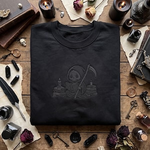 Peut inclure: Un sweat-shirt noir à col rond présentant une broderie gris foncé représentant un personnage de faucheuse à capuche assis lisant un livre avec une faux, entouré de piles de livres et de bougies allumées de chaque côté de la silhouette.