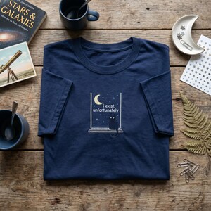May include: A navy blue short-sleeve t-shirt featuring an embroidered graphic of a black cat sitting on a windowsill under a crescent moon and stars. The text "i exist, unfortunately" is embroidered in white letters above the cat.