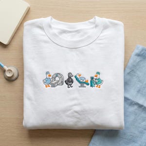 Könnte beinhalten: Weißes Sweatshirt mit Rundhalsausschnitt und einem gestickten Design mit Cartoon-Enten in medizinischen Szenarien. Die Enten werden mit medizinischen Geräten dargestellt, darunter ein MRT-Gerät, ein Röntgenbild und ein Krankenhausbett. Das Design ist in Blau-, Orange- und Schwarztönen gehalten.