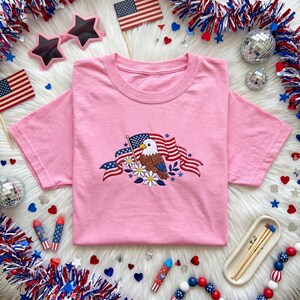 Könnte beinhalten: Ein hellrosa T-Shirt mit kurzen Ärmeln und einer zentralen Stickerei, die einen Weißkopfseeadler neben einer wehenden amerikanischen Flagge zeigt, verziert mit weißen Gänseblümchen und kleinen blauen Sternen auf der Brust.