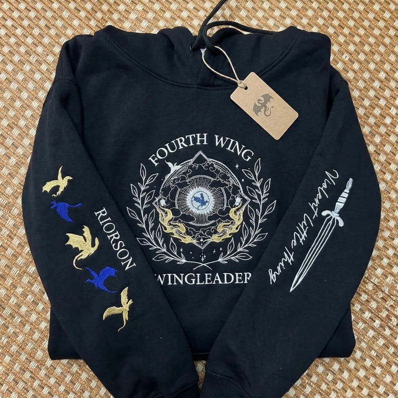May include: Black hooded sweatshirt with embroidered designs. Features the text "FOURTH WING" and "Violent Little Thing". The left sleeve has embroidered dragons and the name "RIORSON". Includes a hang tag with a dragon logo.