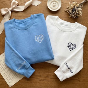 May include: Two crewneck sweatshirts, one light blue and one white, each with a heart-shaped monogram and "Custom text" on the sleeve. The light blue sweatshirt has a white monogram, and the white sweatshirt has a dark blue monogram.