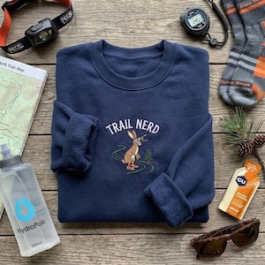 May include: A navy blue crewneck sweatshirt featuring an embroidered graphic of a brown rabbit wearing a headlamp and trail shoes, with the text "TRAIL NERD" arched above. The design includes a winding green trail and a small green pine tree.