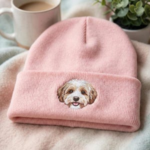 May include: A light pink beanie with a folded brim. The beanie features an embroidered image of a dog's face. The dog has brown and white fur, with its tongue sticking out. The beanie is made of a soft, knitted material.