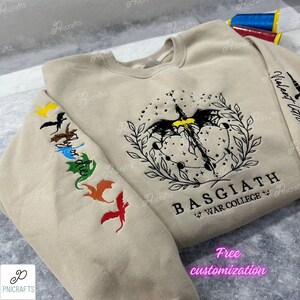 K&ouml;nnte beinhalten: Ein beiger Pullover mit einem schwarzen gestickten Drachenmotiv und dem Text "BASGIATH WAR COLLEGE". Der Pullover hat bunte gestickte Drachen auf dem &Auml;rmel.