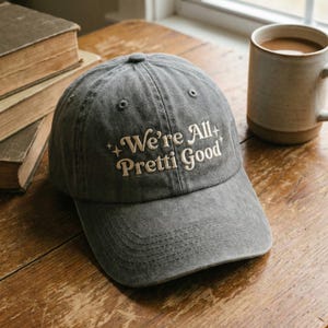 May include: A grey baseball cap with the embroidered text "We're All Pretti Good" in cream. The hat is on a wooden table, beside a mug of coffee and a stack of books.