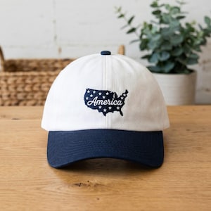 May include: White baseball cap with a navy blue peak and button. The cap features an embroidered design of the United States map with stars and the word "America". A stylish accessory.