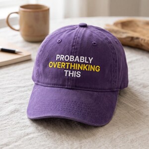 Peut inclure: Une casquette de baseball violette avec le texte brodé PROBABLY OVERTHINKING THIS en lettres blanches et jaunes. La casquette présente une calotte structurée, une visière incurvée et des œillets cousus sur les panneaux supérieurs pour la ventilation.