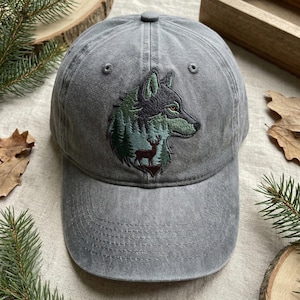 May include: Grey baseball cap featuring an embroidered design of a wolf's head, forest, and deer. The wolf is black and green, with a red eye. The forest is green and blue. The cap has a curved peak and a button on top.