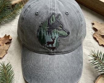 May include: A grey baseball cap featuring an embroidered design of a wolf head silhouette filled with a forest scene and a small deer standing at the base. The cap has a curved brim, a button on top, and stitched eyelets for ventilation.