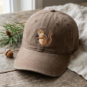 Peut inclure: Une casquette de baseball marron avec une broderie représentant un tamia tenant un gland. La casquette présente une calotte structurée avec des œillets d'aération et une visière incurvée, fabriquée dans un tissu uni avec des coutures visibles.