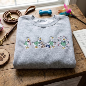 May include: A folded gray sweatshirt with an embroidered design of cartoon ducks in medical attire caring for pets. The ducks are depicted with a dog, a cat, and paw prints. A dog leash, toy, and notepad are in the background.