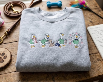 May include: A folded gray sweatshirt with an embroidered design of cartoon ducks in medical attire caring for pets. The ducks are depicted with a dog, a cat, and paw prints. A dog leash, toy, and notepad are in the background.
