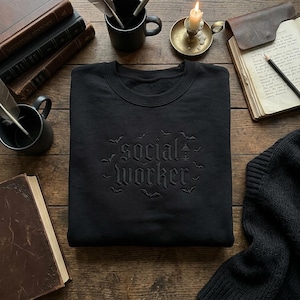 May include: A black crewneck sweatshirt featuring dark, tone-on-tone embroidery on the chest. The text reads "social worker" in a gothic-style font, surrounded by small embroidered bat silhouettes and a single lantern icon.