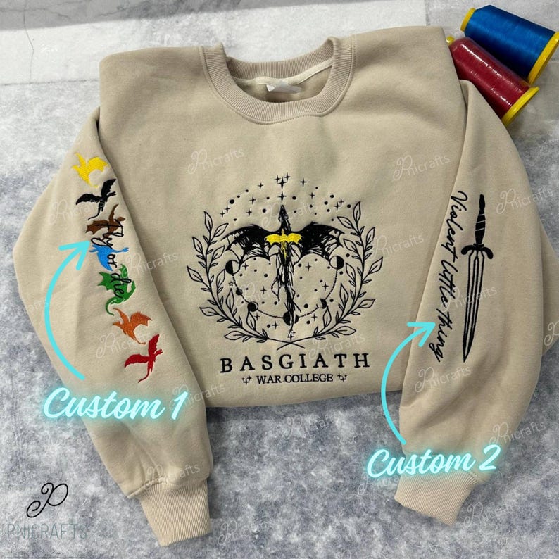 K&ouml;nnte beinhalten: Ein beiger Pullover mit zwei verschiedenen Stickereien auf den &Auml;rmeln. Ein &Auml;rmel hat ein Design mit einem Schwert mit dem Text "Violent little thing" und der andere &Auml;rmel hat ein Design mit f&uuml;nf bunten Drachen. Die Vorderseite des Pullovers hat ein Schwarzwei&szlig;-Design eines Drachen mit dem Text "BASGIATH WAR COLLEGE".