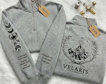 May include: Two gray quarter-zip sweatshirts. One has a sleeve design with moon phases and the text "To the stars Who listen and the dreams that are answered." The other has a mountain design with the text "VELARIS CITY OF STARLIGHT."