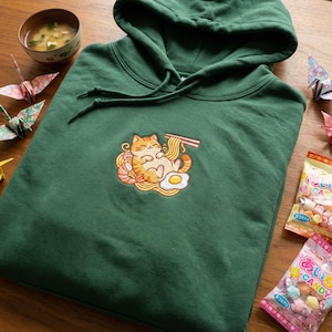 May include: A dark green hoodie with a drawstring hood and a front pocket. The hoodie features an embroidered design of a cartoon cat enjoying ramen, shrimp, and a fried egg. Origami cranes and candy are in the background.
