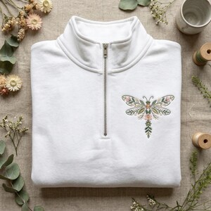 May include: White quarter-zip sweatshirt with a floral dragonfly embroidery. The dragonfly design features pink and green flowers and leaves. The sweatshirt has a silver zipper and a high collar.