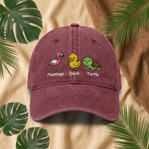 May include: A maroon baseball cap with cartoon images of a flamingo, duck, and turtle. The text below the images reads "Flamingo", "Duck", and "Turtle". The cap has a curved peak and an adjustable strap.