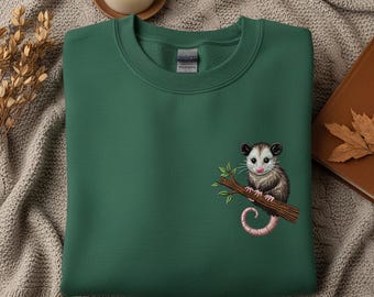 May include: A dark green sweatshirt featuring an embroidered opossum design. The opossum is perched on a branch with green leaves and has a pink tail. The sweatshirt is made of a soft material.