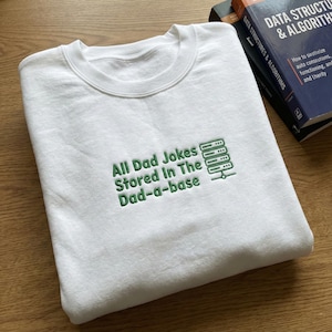 May include: White sweatshirt with the embroidered text "All Dad Jokes Stored In The Dad-a-base" in green. The design includes a server rack graphic. Two books are visible in the background.