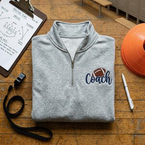May include: A grey quarter-zip sweatshirt featuring the word "Coach" embroidered in navy blue with a football graphic. The sweatshirt has a high collar and a silver zip. A clipboard with a practice plan and a whistle are also visible.