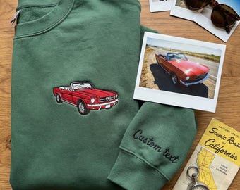 May include: A dark green sweatshirt featuring a red convertible car embroidery. The sleeve cuff has the words "Custom text" embroidered in black. The image also includes sunglasses, photos, and a vintage travel brochure.