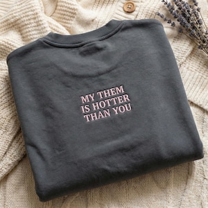 May include: A dark gray sweatshirt with the embroidered text "MY THEM IS HOTTER THAN YOU" in pink. The sweatshirt is folded on a cream-colored knitted surface, with a sprig of lavender and a button visible.