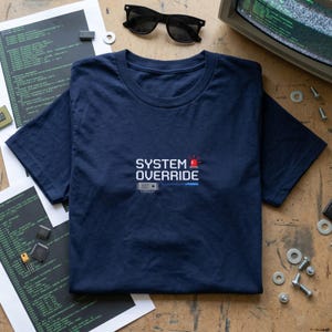 May include: Navy blue t-shirt with the text "SYSTEM OVERRIDE" in white, a red alarm icon, and a blue line graphic. The shirt is on a wooden surface with computer components, code printouts, and sunglasses.