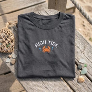 May include: Charcoal grey t-shirt featuring the embroidered words "HIGH TIDE" above an orange crab design. The shirt is folded on a wooden surface, evoking a relaxed, seaside aesthetic.
