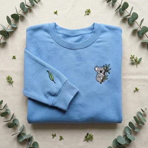 May include: A pale blue sweatshirt with a crew neck and long sleeves. It has an embroidered koala holding a branch on the front and a single leaf on the left sleeve. The background is a neutral colour with eucalyptus leaves.