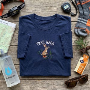 May include: Navy blue t-shirt with the text "TRAIL NERD" and an embroidered rabbit design. The image also includes a headlamp, a trail map, a water bottle, a stopwatch, socks, sunglasses, and a GU energy gel.