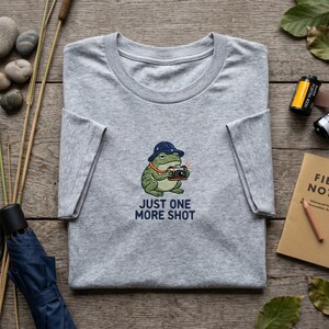 Könnte beinhalten: Ein meliertes graues T-Shirt mit einer Stickerei einer Froschfigur mit blauem Hut und Kamera. Der Text "JUST ONE MORE SHOT" steht darunter. Das T-Shirt liegt gefaltet auf einer Holzoberfläche.