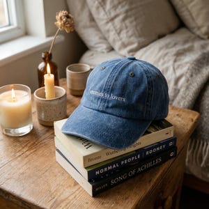 May include: A blue denim baseball cap with the phrase "enemies to lovers" embroidered in white. The cap rests on a stack of books on a wooden table, next to lit candles and a small vase. The books are titled "Wuthering Heights", "Normal People", and "Song of Achilles".
