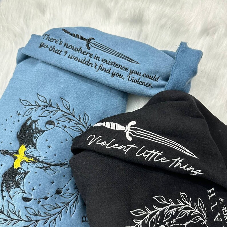 May include: Two hooded sweatshirts with embroidered designs. The blue sweatshirt has a moon and stars design with the text "There's nowhere in existence you could go that I wouldn't find you, Violence". The black sweatshirt has a sword design with the text "Violent little thing".